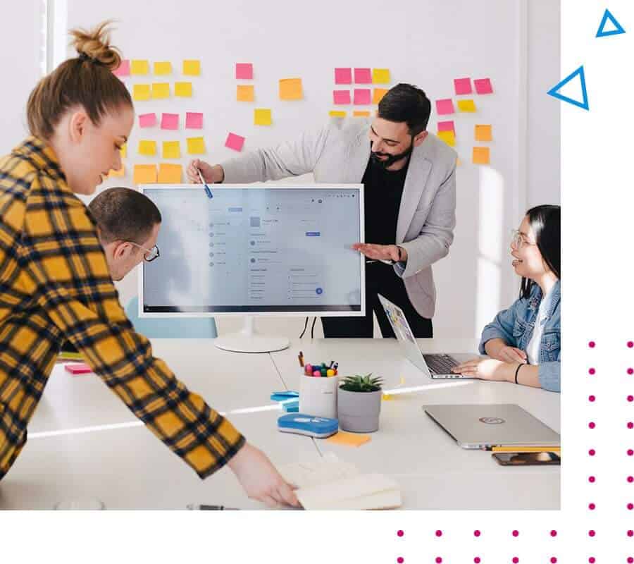 Das Bild zeigt vier Personen in einem hellen Büro, die offenbar in einer Geschäfts- oder Arbeitsbesprechung mit Post-its an der Wand und einem großen Bildschirm sind.