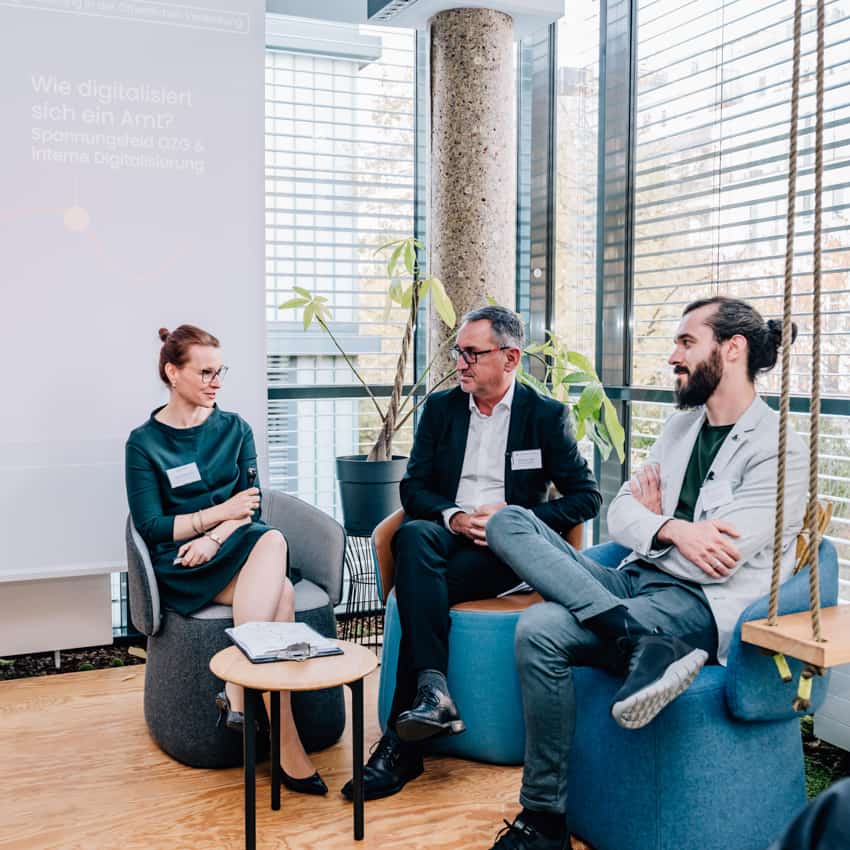 Drei Personen sitzen und scheinen an einer Diskussion oder einem Gesprächsforum teilzunehmen, möglicherweise im Rahmen einer Konferenz oder eines Meetings mit dem Thema Digitalisierung in der Verwaltung, wie es der Text auf dem Banner andeutet.