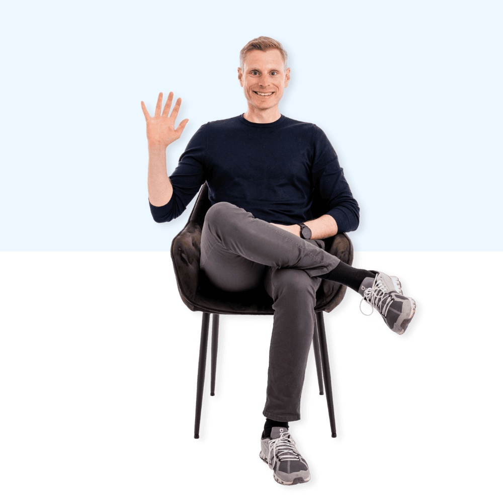 Auf dem Bild ist ein lächelnder Mann zu sehen, der auf einem Stuhl sitzt und mit einer Hand winkt.
