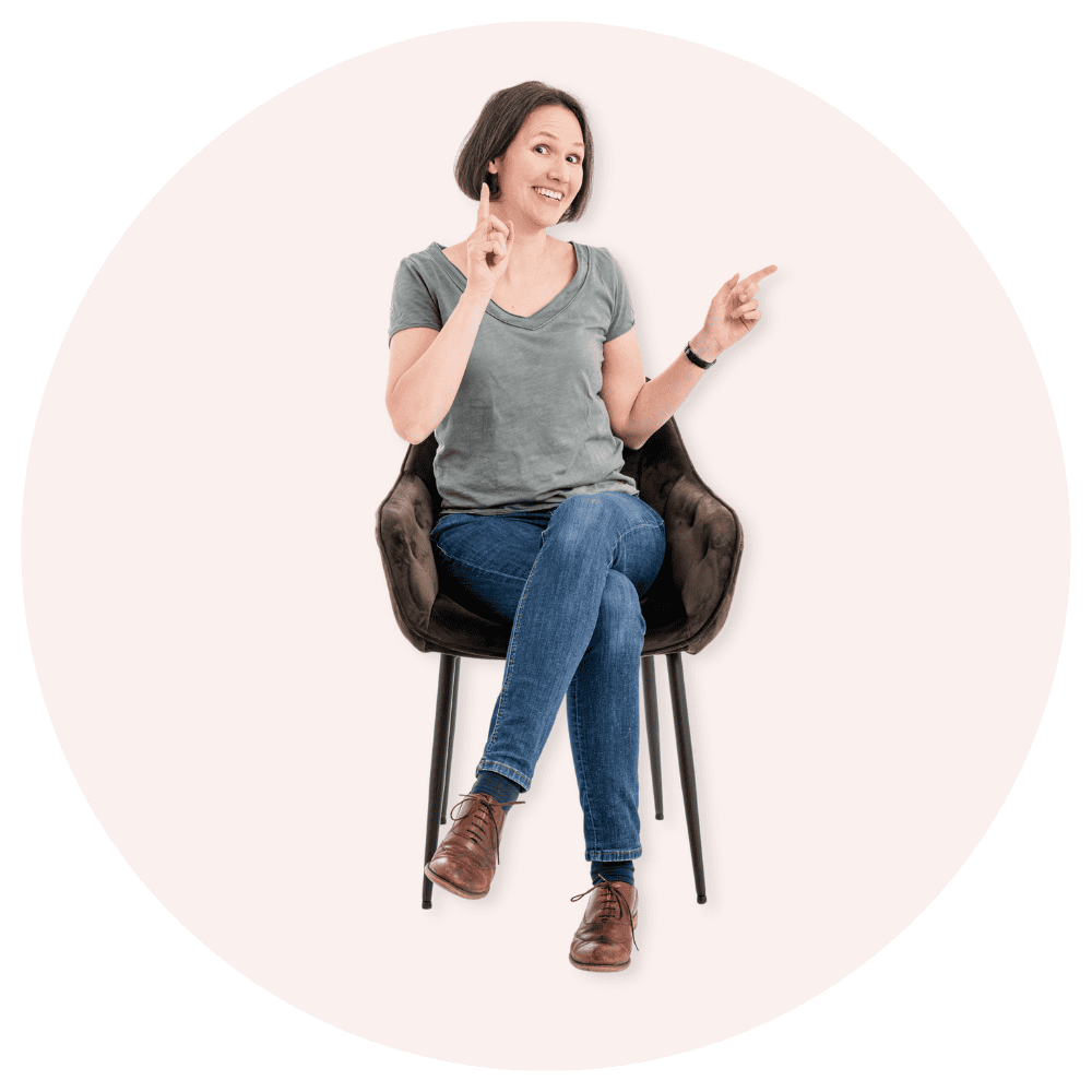 Auf dem Bild ist eine lächelnde Frau, die sitzt und mit ihrer Hand etwas andeutet.