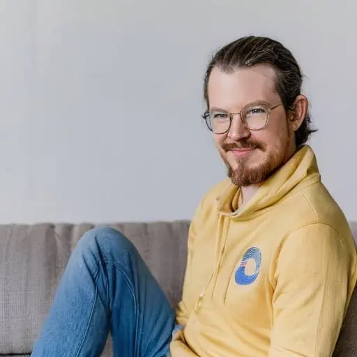 Auf dem Bild ist eine Person mit Brille, die auf einem Sofa sitzt und einen gelben Kapuzenpullover sowie blaue Jeans trägt.