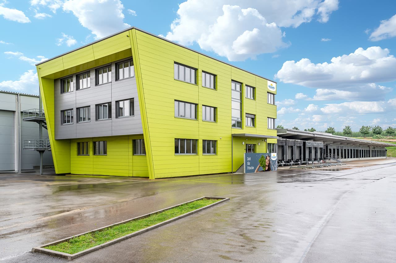 Das ist ein modernes Gebäude in leuchtendem Gelb neben einer Laderampe oder Logistikzone mit bewölktem Himmel im Hintergrund.