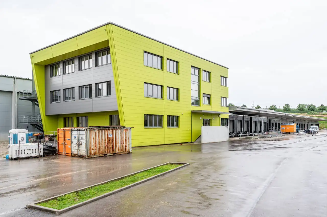Das Bild zeigt ein modernes Gebäude mit hellgelber Fassade und grauen Elementen auf einem Industrie- oder Gewerbegelände bei nasser Witterung.