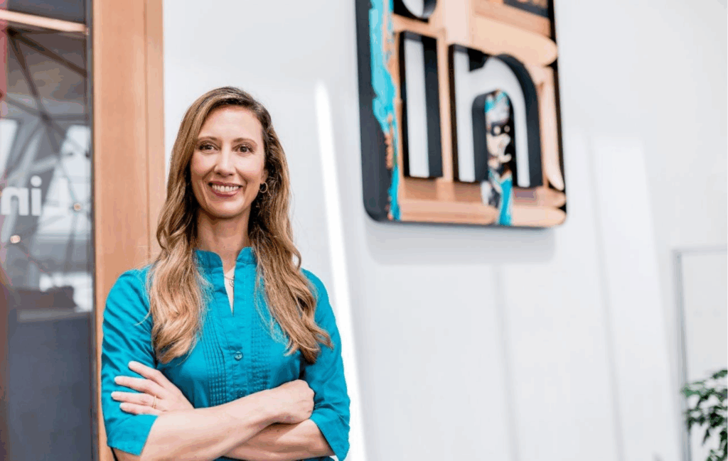Das Bild zeigt eine selbstbewusste Frau in einer modernen Büroumgebung, die mit ihrem freundlichen Lächeln und ihrem blauen Oberteil für Professionalität und Zugänglichkeit steht. Der Hintergrund mit dem markanten "in"-Logo betont das kreative und innovative Arbeitsumfeld, was es zu einem idealen Symbol für Unternehmertum und Networking macht.