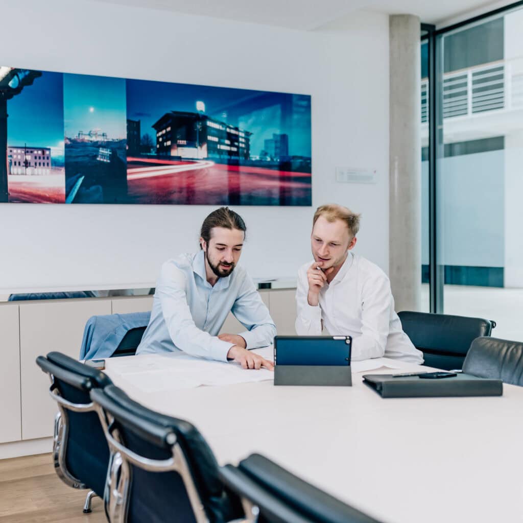 In einem modernen Meetingraum analysieren zwei Fachleute gemeinsam digitale Informationen auf einem Tablet, während sie an einem großen Tisch sitzen, der mit Dokumenten und Notizen bedeckt ist. Die stilvolle Umgebung und das kreative Wandbild an der Rückwand fördern eine inspirierende Arbeitsatmosphäre für effiziente Teamarbeit und innovative Lösungen.
