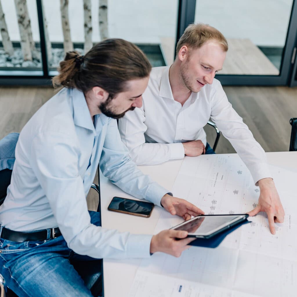 In einem modernen Büro-Setting arbeiten zwei Fachleute konzentriert an einem Projekt, während sie Pläne und digitale Geräte analysieren. Diese Zusammenarbeit symbolisiert effizientes Projektmanagement und innovative Lösungsfindung, die entscheidend für den Geschäftserfolg sind.