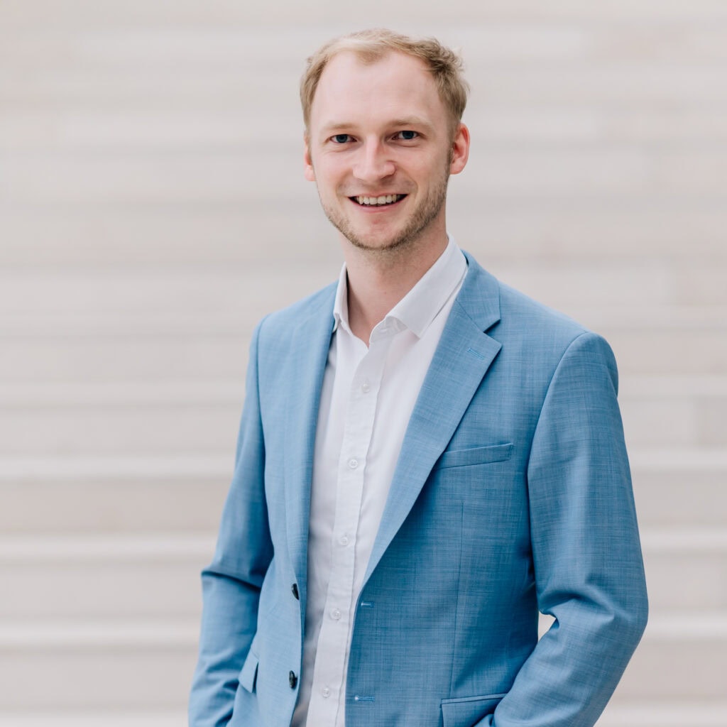 Der junge Mann in einem stilvollen blauen Anzug strahlt Selbstbewusstsein und Professionalität aus und lächelt charmant in die Kamera. Die helle, minimalistische Umgebung mit sanften Treppen im Hintergrund verstärkt den eleganten und modernen Eindruck des Porträts, ideal für berufliche Netzwerke oder persönliche Branding-Zwecke.