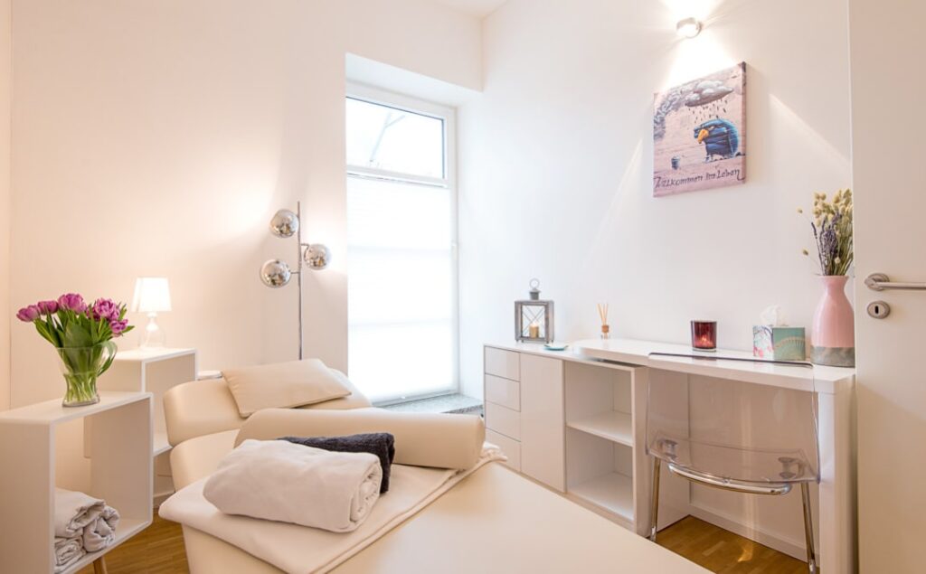 Das Bild zeigt einen modernen, hellen Raum mit einem einladenden Ambiente, ideal für Entspannung und Wellness. Die stilvolle Einrichtung, einschließlich einer gemütlichen Liege, frischen Blumen und sanfter Beleuchtung, schafft eine harmonische Atmosphäre für Behandlungen und Erholung.