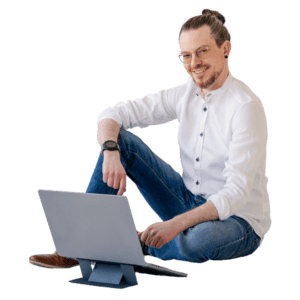 Ein selbstbewusster junger Mann sitzt entspannt am Boden und arbeitet an seinem Laptop, was eine moderne Arbeitsumgebung symbolisiert. Sein legerer Stil mit einem weißem Hemd und Jeans strahlt Kreativität und Flexibilität aus, ideal für Themen wie Remote-Arbeit und digitales Nomadentum.