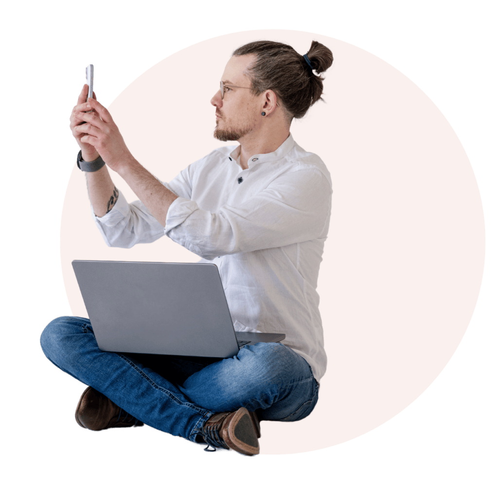 Der junge Mann arbeitet kreativ und flexibel, während er mit seinem Smartphone interagiert und sich auf seinem Laptop konzentriert. Diese Szene verkörpert modernes Arbeiten und digitales Nomadentum, ideal für Themen wie Homeoffice, digitale Kommunikation und moderne Arbeitsweisen.