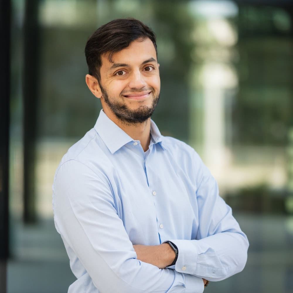 Das Bild zeigt einen selbstbewussten Mann in einem hellblauen Hemd, der mit verschränkten Armen vor einer modernen, architektonischen Kulisse steht. Diese professionelle Darstellung eignet sich ideal für Themen wie Business, Personal Branding und Karriereentwicklung.