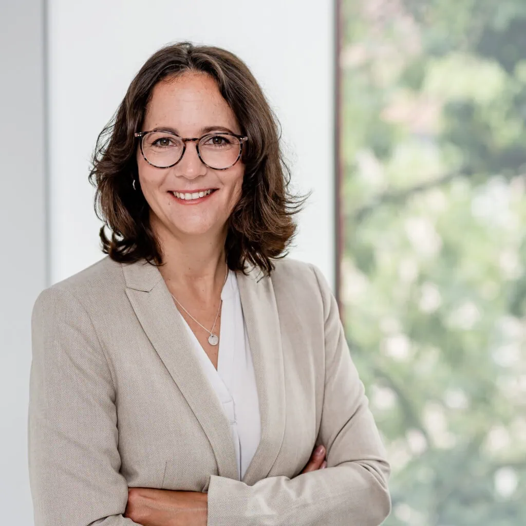 Das Bild zeigt eine selbstbewusste Frau in einem eleganten Business-Anzug, die mit einem freundlichen Lächeln in die Kamera blickt. Der Hintergrund aus sanften Farben und natürlichen Elementen vermittelt eine professionelle und einladende Atmosphäre, ideal für Unternehmenspräsentationen oder Karriereprofile.