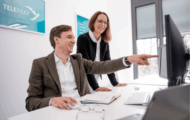 Das Bild zeigt einen dynamischen Austausch zwischen zwei Fachleuten in einem modernen Büro, in dem sie gemeinsam an einem Computer arbeiten. Die freundliche Atmosphäre und das Sichtfeld auf digitale Lösungen spiegeln die Innovationskraft und Teamarbeit von TELEPAXX wider, einem wertvollen Partner für Kliniken und Radiologie.